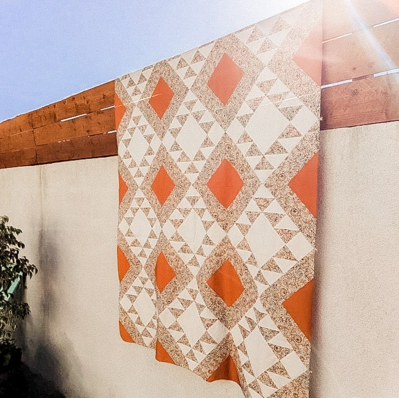 May include: A white and orange quilt with a geometric pattern. The quilt is hanging on a wall.