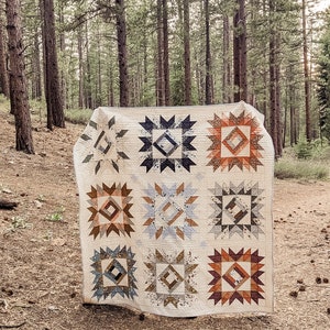 May include: A white quilt with a geometric pattern in shades of brown, blue, and orange. The quilt is displayed in a forest setting.