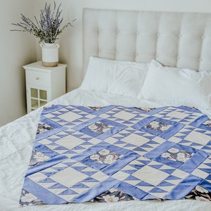 May include: A white bed with a blue and white patterned quilt. The bed has a white headboard with buttons. There is a vase of lavender flowers on a white nightstand next to the bed.