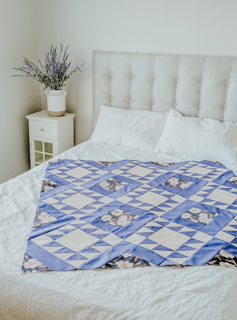 May include: A white bed with a blue and white patterned quilt. The bed has a white headboard with buttons. There is a vase of lavender flowers on a white nightstand next to the bed.