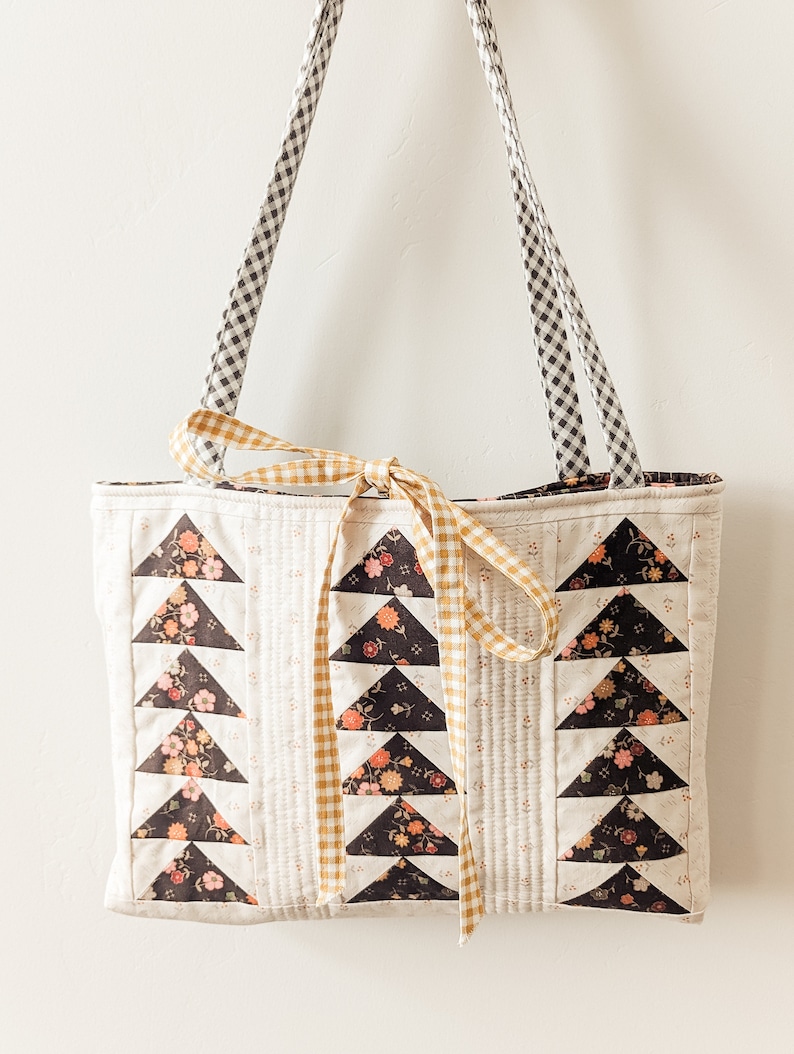Peut inclure: Un sac cabas matelass&eacute; blanc avec des triangles en tissu noir et floral. Le sac est dot&eacute; d'un ruban jaune et blanc &agrave; carreaux nou&eacute; autour des poign&eacute;es et de sangles en gingham noir et blanc.