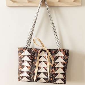 Peut inclure: Un sac cabas matelass&eacute; marron avec des motifs triangulaires blancs et un ruban en vichy jaune. Le sac est suspendu &agrave; un porte-manteau en bois avec des crochets.