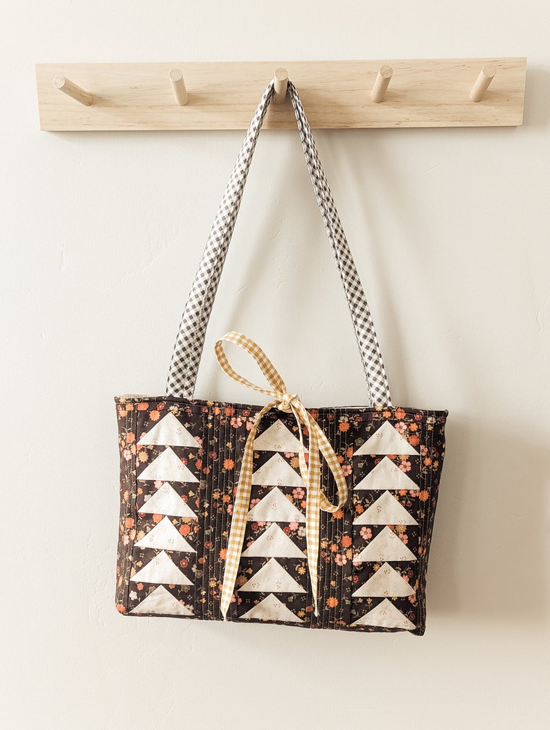 Peut inclure: Un sac cabas matelass&eacute; marron avec des motifs triangulaires blancs et un ruban en vichy jaune. Le sac est suspendu &agrave; un porte-manteau en bois avec des crochets.