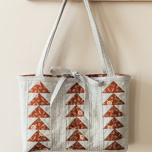 Peut inclure: Un sac cabas gris avec des triangles en tissu floral brun et blanc sur le devant. Le sac a une longue bandouli&egrave;re grise et un n&oelig;ud nou&eacute; en haut.
