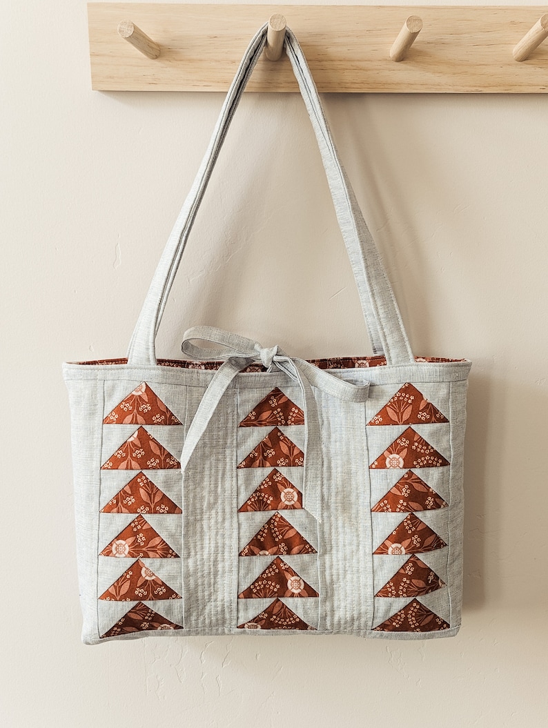 Peut inclure: Un sac cabas gris avec des triangles en tissu floral brun et blanc sur le devant. Le sac a une longue bandouli&egrave;re grise et un n&oelig;ud nou&eacute; en haut.