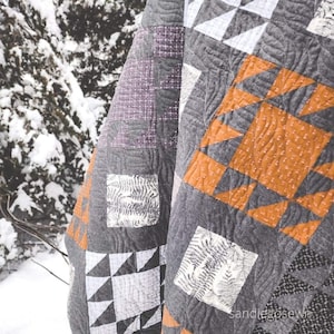 May include: A gray and white quilt with a geometric pattern. The quilt is made of different fabrics and has a variety of textures. The quilt is folded and draped over a snowy branch.