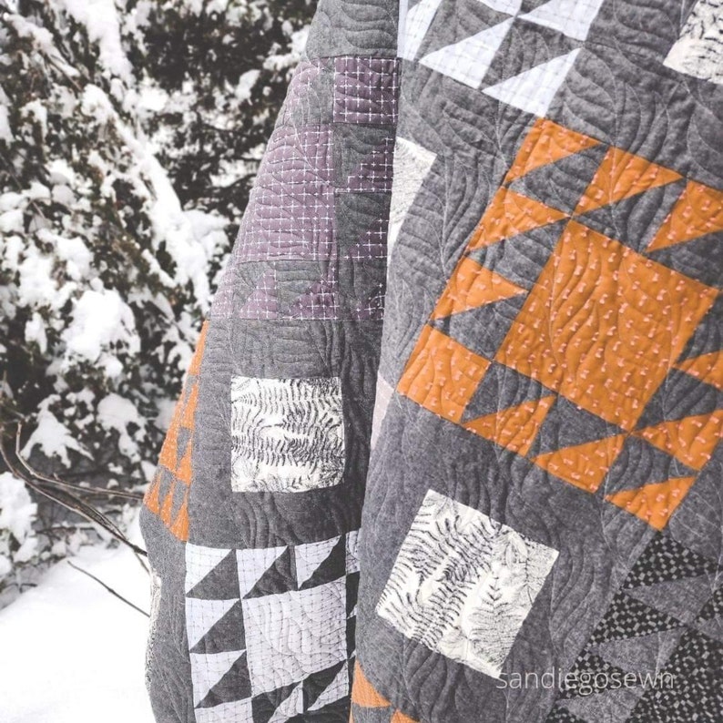 May include: A gray and white quilt with a geometric pattern. The quilt is made of different fabrics and has a variety of textures. The quilt is folded and draped over a snowy branch.