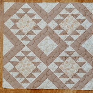 May include: A beige and white quilt with a geometric pattern. The quilt is made of squares and triangles, and the pattern is repeated throughout the quilt.