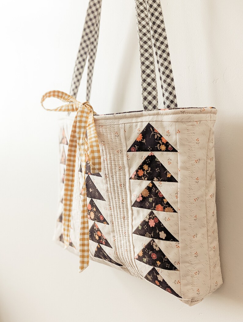 Peut inclure: Un sac cabas matelass&eacute; blanc avec des triangles en tissu &agrave; motifs floraux noirs. Le sac a deux anses en vichy noir et blanc et un ruban de nouage en vichy jaune.