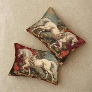 Op de afbeelding: Twee decoratieve sierkussens met een vintage equestrian design. Elk kussen heeft een wit paard met bloemaccenten tegen een achtergrond van een bossc&egrave;ne met rode draperie&euml;n. De kussens zijn ruitvormig.