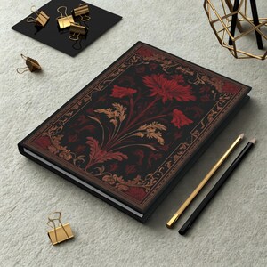 Op de afbeelding: Een zwart notitieboek met een bloemmotief in rood en goud. Het notitieboek is omgeven door gouden paperclips, potloden en een zwart vierkant. De omslag heeft een gedetailleerde botanische illustratie.