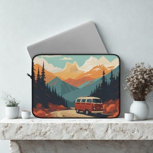 May include: Laptop sleeve featuring a retro-style illustration of a red and white van driving on a winding road through a mountain landscape. The design includes pine trees, mountains, and a teal and orange color palette.