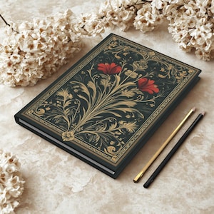 Op de afbeelding: Een hardcover notitieboek met een donkerblauwe omslag met een gouden bloemenontwerp en twee rode bloemen. Twee potloden, &eacute;&eacute;n goud en &eacute;&eacute;n zwart, liggen naast het notitieboek. Het notitieboek ligt op een lichtbeige oppervlak.