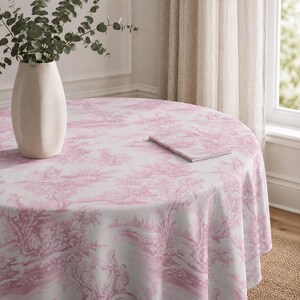 May include: A round tablecloth with a pink and white toile pattern featuring a pastoral scene. A matching notebook rests on the table. A beige vase with eucalyptus branches sits on the table. The tablecloth is draped over a round table.
