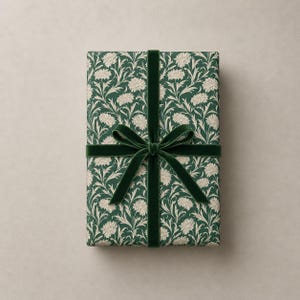 K&ouml;nnte beinhalten: Ein rechteckiges Geschenk, verpackt in dunkelgr&uuml;nem Papier mit wei&szlig;em Blumenmuster, gebunden mit einem dunkelgr&uuml;nen Samtband und einer Schleife. Das Papier hat ein sich wiederholendes Muster aus wei&szlig;en Blumen und Bl&auml;ttern.