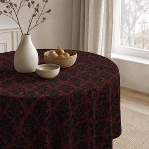 May include: A round tablecloth with a dark red and black floral pattern covers a table. A white vase with branches, a small bowl, and a basket of pears sit on the table. The table is near a window with a beige curtain.