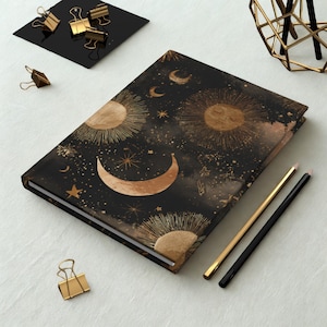 May include: A black journal with a celestial design featuring gold suns, moons, and stars. Gold pencils and binder clips are next to the journal. The journal's cover has a detailed, cosmic pattern.