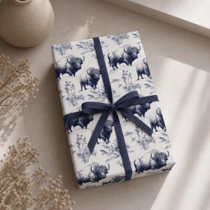 Op de afbeelding: Een ingepakt cadeau met wit papier met een herhalend patroon van donkerblauwe bizons en bladeren. Het cadeau is vastgebonden met een donkerblauw lint en strik. Een beige vaas en droogbloemen staan op de achtergrond.