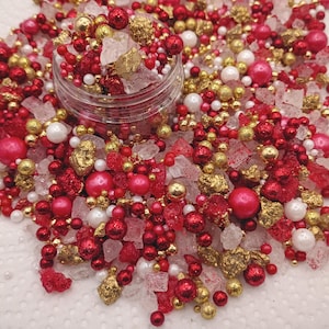May include: A clear plastic jar filled with a decorative mix of red, gold, and white sprinkles. The assortment includes spherical beads in various sizes, metallic gold nuggets, and translucent white sugar crystals, creating a textured blend.