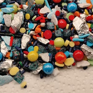 May include: A colorful assortment of sprinkles, including blue, red, yellow, and orange spheres, silver-colored shards, and small, dark blue rod-shaped sprinkles. The sprinkles are scattered on a white, textured surface.