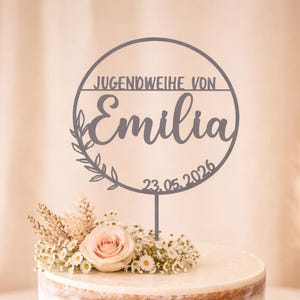 Könnte beinhalten: Eine zweistöckige Torte auf einem Holzständer, verziert mit frischen Blumen und einem Tortenaufsatz. Der Tortenaufsatz lautet "JUGENDWEIHE VON Emilia 23.05.2026". Die Torte ist mit rosa Rosen und kleinen weißen Blumen dekoriert.
