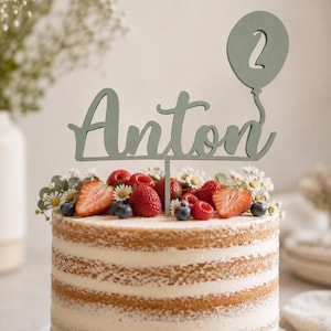 Könnte beinhalten: Ein salbeigrüner Tortenstecker mit dem Namen Anton in Schreibschrift und einem Ballon-Motiv, das die Zahl 2 enthält. Der Tortenstecker steckt in einer runden Torte, die mit frischen Erdbeeren und Blaubeeren verziert ist.