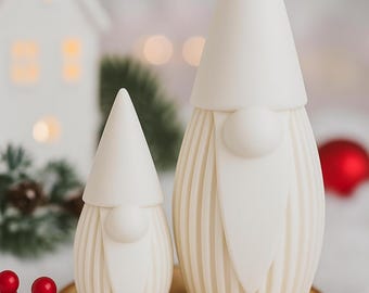 May include: Two white gnome figurines with conical hats and long beards. The gnomes are placed on a wooden slice, with a small white house and red ornaments in the background. The larger gnome is approximately 6 inches tall.