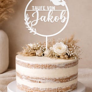 Op de afbeelding: Een witte taart met een decoratieve taarttopper met de tekst "Taufe von Jakob" en een duif. De taart is versierd met een bloemstuk van witte rozen, droogbloemen en kleine eieren. De taart staat op een witte taartstandaard.