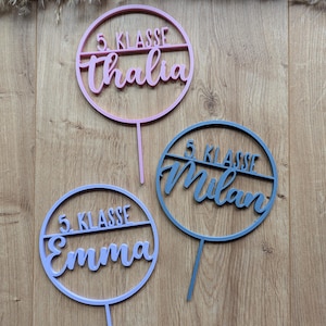 Könnte beinhalten: Drei kreisförmige Tortenstecker mit einem horizontalen Mittelsteg. Jeder Stecker zeigt den Text 5. KLASSE über einem Namen in Schreibschrift: thalia in Pink, Milan in Dunkelblau und Emma in Helllila. Jeder Stecker hat einen vertikalen Stiel.