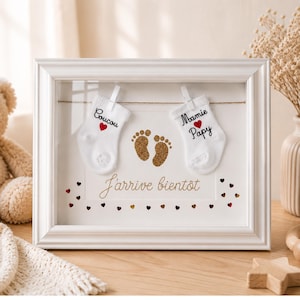 Peut inclure: Un cadre blanc pour bébé avec deux petites chaussettes blanches, chacune avec du texte et un cœur rouge. Le cadre présente également des empreintes de pieds en paillettes dorées et les mots "J'arrive bientôt".