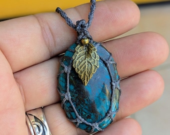 May include: An oval-shaped blue and dark brown stone pendant encased in a grey macrame cord net. The pendant features a small metallic gold-toned leaf charm attached at the top, suspended from a matching grey cord necklace.