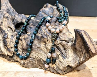 May include: A green and pink gemstone mala necklace with a decorative metal charm. The necklace is displayed on a piece of driftwood.