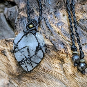 May include: A clear quartz pendant with black inclusions, encased in a macrame net, hangs from a black cord. The necklace features small beads and a gold-colored accent. The pendant is displayed on a textured, wooden surface.