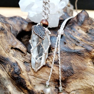 Puede incluir: Un colgante de cristal de cuarzo transparente envuelto en cordón de macramé con un pequeño dije de hoja. El colgante cuelga de un cordón beige con dos cuentas al final.