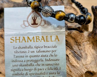 Puede incluir: Un brazalete de cordón negro con cuentas de ojo de tigre, cuentas negras y un dije de plata. El brazalete está sobre una superficie de madera con una tarjeta que dice "SHAMBALLA" y describe el brazalete como un talismán tibetano para el alma.