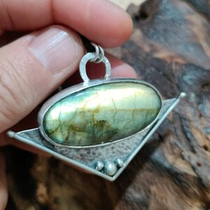 Puede incluir: Un colgante de plata con una piedra preciosa de labradorita verde y dorada engastada en un diseño geométrico.