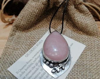 May include: A pink quartz pendant necklace with a silver setting featuring a leaf design. The pendant is on a black cord and is displayed on a burlap bag. The text on the card reads: "Il Quarzo Rosa, considerata della fertilità, rafforzando se stessi che gli aumenta l'amore dell'anima. Sta disponibilità confronti dell' funzione car l'irrorazione sa una"