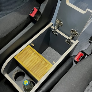Puede incluir: Una consola de coche de madera hecha a medida con una tapa con bisagras, un peque&ntilde;o estante y dos portavasos. La consola est&aacute; instalada en el &aacute;rea de la consola central de un coche, entre los asientos del conductor y del pasajero.