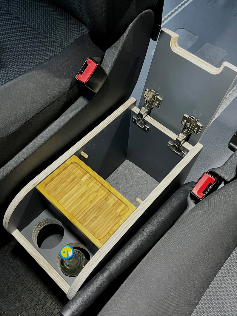 Puede incluir: Una consola de coche de madera hecha a medida con una tapa con bisagras, un peque&ntilde;o estante y dos portavasos. La consola est&aacute; instalada en el &aacute;rea de la consola central de un coche, entre los asientos del conductor y del pasajero.