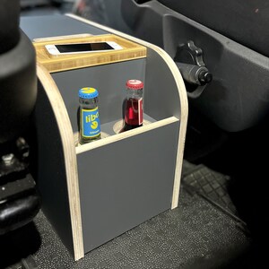 Puede incluir: Un organizador de coche de madera con un interior gris y dos ranuras para botellas. El organizador tiene una tapa de bamb&uacute; con un soporte para tel&eacute;fono. El organizador est&aacute; en un coche con una alfombra de suelo negra.