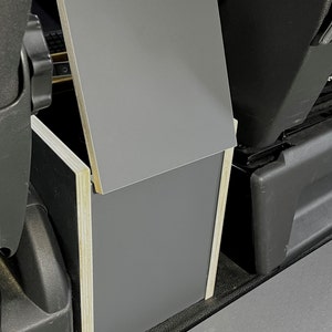 Puede incluir: Una caja de almacenamiento gris con una tapa con bisagras, hecha de madera y contrachapado, est&aacute; instalada en un veh&iacute;culo. La caja est&aacute; abierta y la tapa est&aacute; levantada.