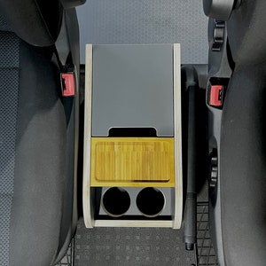 Puede incluir: Un organizador de consola de coche de madera con una parte superior gris, un estante de bamb&uacute; y dos portavasos. El organizador est&aacute; instalado entre dos asientos de coche.