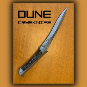 以下が含まれることがあります： DUNEのCrysknifeのレプリカ。長く尖った銀色の刃、柄の近くに複雑なエッチング模様、暗い質感のハンドルが特徴。刃の上部には、黒く太いサンセリフ体の大文字で「DUNE CRYSKNIFE」という文字が印刷されている。