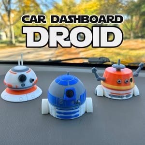 May include: Three small droid figurines displayed on a car dashboard. The left droid is white and orange, the center droid is blue and white, and the right droid is orange and white with raised arms. Text above reads CAR DASHBOARD DROID.