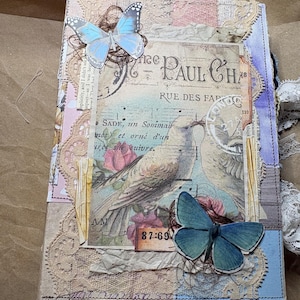 May include: A rectangular journal with a collage cover featuring two blue butterfly cutouts, a central image of a bird, vintage French text including Paul Cha, musical notes, and layered beige lace accents. The spine has white lace trim.