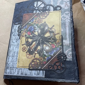 May include: Black leather journal with a steampunk design. The cover features a metal octopus with gears and a clock face. The journal has a black string attached to it.