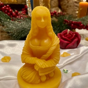 May include: A yellow beeswax candle sculpted in the likeness of a woman wearing a shawl. The candle is sitting on a yellow base.
