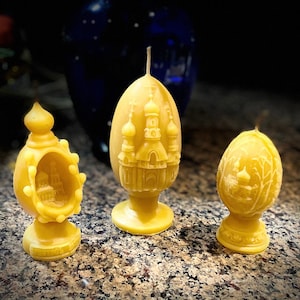May include: Three yellow beeswax candles shaped like eggs with intricate carvings of churches and trees. The candles are on a brown surface.