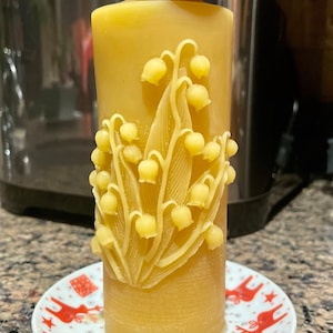 May include: A yellow beeswax candle with a lily of the valley design. The candle is sitting on a white saucer with a red reindeer pattern.