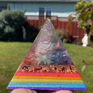 May include: A colorful pyramid-shaped resin art piece. The pyramid features layers of rainbow colors, gold flakes, and crystals. The top of the pyramid has a clear crystal wrapped in copper wire. The pyramid is held in a hand.